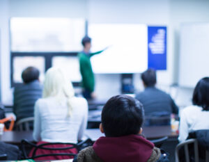 far-focused image of a teacher at a display board with the backs of students' heads in the foreground