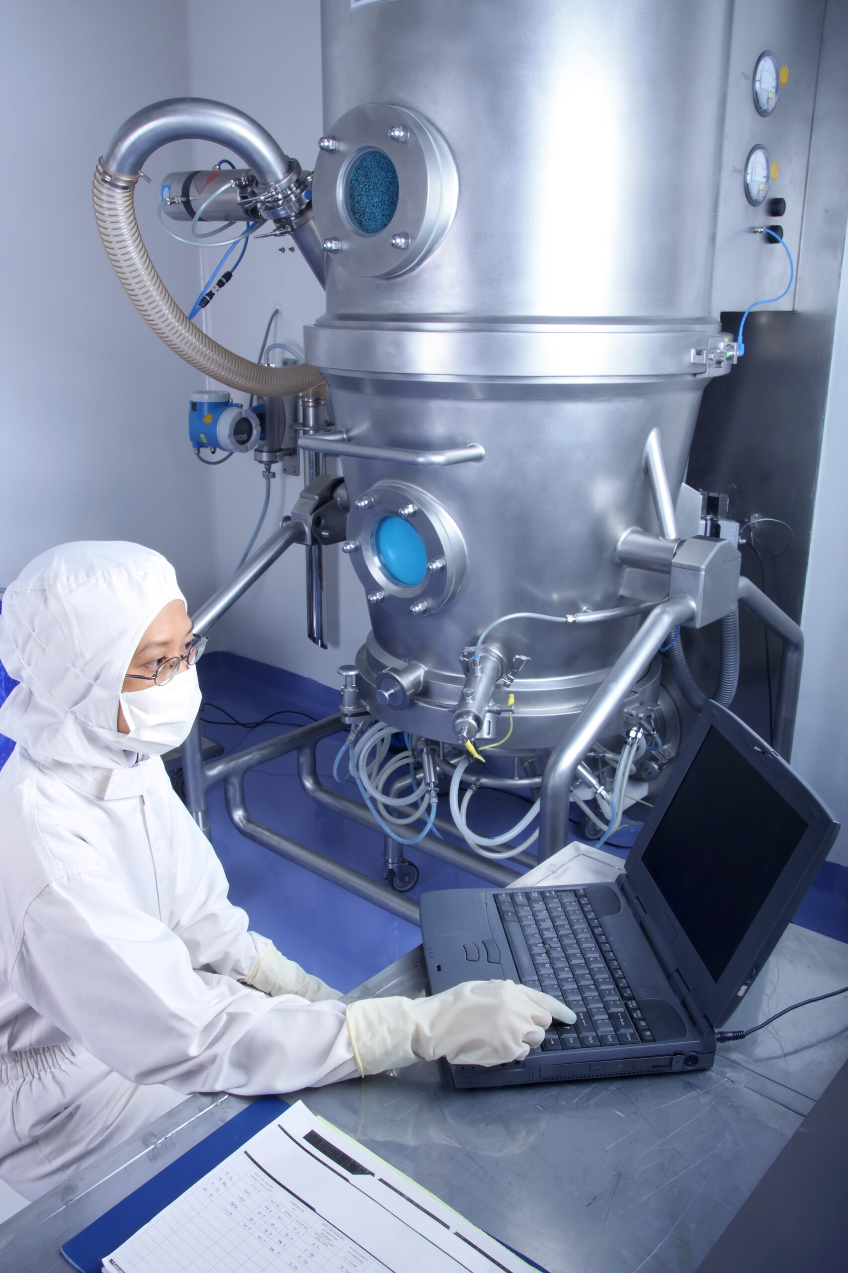 A gowned technician sits at a laptop computer next to a large piece of manufacturing equipment